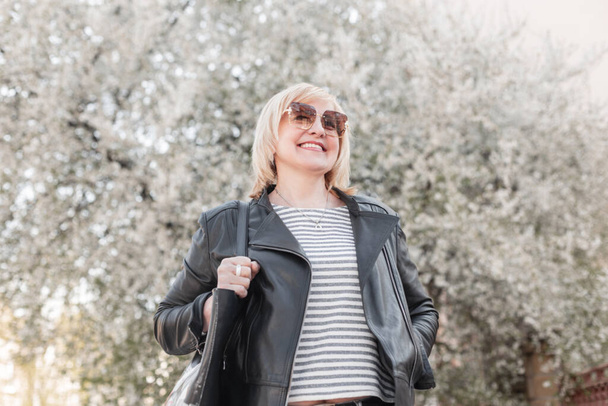 Beautiful elderly happy fashionable woman with sunglasses in fashion clothes and a leather jacket and stylish purse walks and enjoy in a park with flowering trees - Photo, image