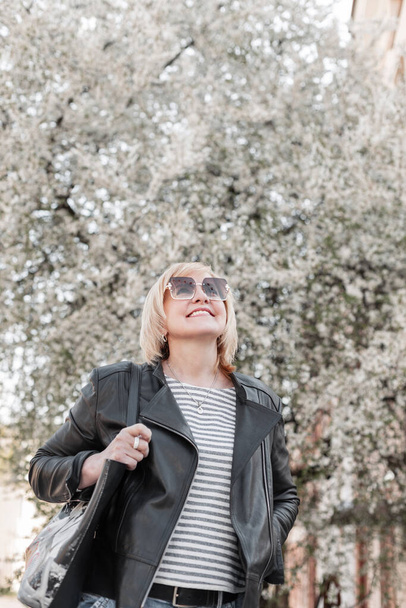 Glücklich stilvolle schöne ältere Frau mit einem Lächeln mit trendiger Sonnenbrille in modischer Kleidung mit Lederjacke und T-Shirt und Handtasche zu Fuß und genießen in der Nähe eines blühenden Parks in der Stadt - Foto, Bild