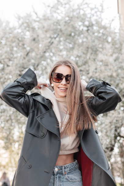 Beautiful young stylish happy girl with a smile in fashionable spring outfit with a long leather coat, hoodie and fashion sunglasses is wearing a hood and walking in the park - Valokuva, kuva