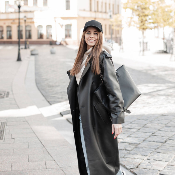 Krásná módní mladá žena model s čepicí ve stylovém černém oblečení vzhled s džínovou koženou bundou a mikina s taškou procházky ve městě za slunečného dne - Fotografie, Obrázek