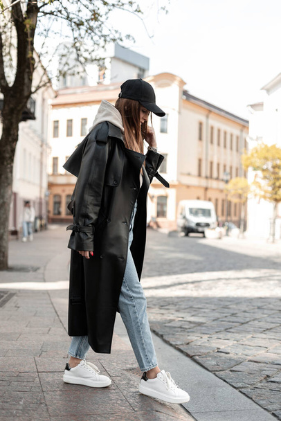 Mode schöne junge Frau Modell Hipster in modischen schwarzen Freizeitkleidung mit einer Kappe in einem langen Ledermantel mit Jeans und weißen Schuhen zu Fuß in der Stadt. Cooler weiblicher urbaner Outfit-Look - Foto, Bild