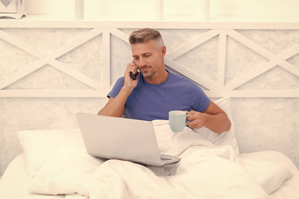 Happy man talk on mobile phone drinking coffee and using laptop in bed in morning, communication. - Foto, imagen