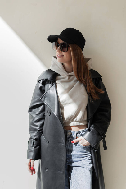 Fashionable beautiful girl with sunglasses in stylish clothes with a cap, hoodie and fashionable leather coat stands near the wall in the sunlight - Fotoğraf, Görsel