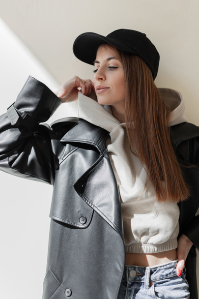 Beautiful trendy young hipster woman with a fashion black mockup cap in a stylish leather coat with a fashionable hoodie stands and poses near a beige wall in the sunlight. Urban female style look - Foto, afbeelding