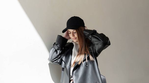 Beautiful young girl in a fashionable black cap with a stylish leather jacket and hoodie walks in the city near the wall with the rays of the sun - 写真・画像