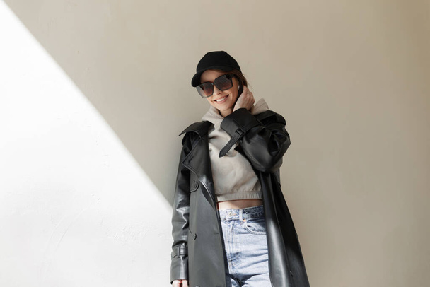 Trendy fashionable happy beautiful young girl with a smile with cool sunglasses and a black cap mockup in a fashion leather coat with a hoodie and jeans stands near the wall in the street with light - Foto, afbeelding