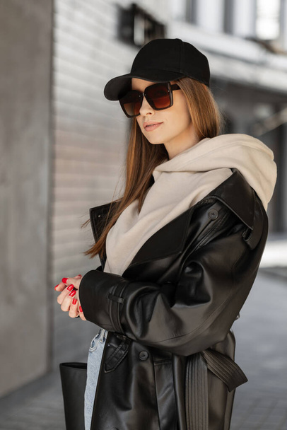 Modieus mooi stijlvol hipster meisje met stijlvolle vintage zonnebril en een zwarte cap mockup in trendy urban kleding met een capuchon, leren jas en tas lopend op straat - Foto, afbeelding