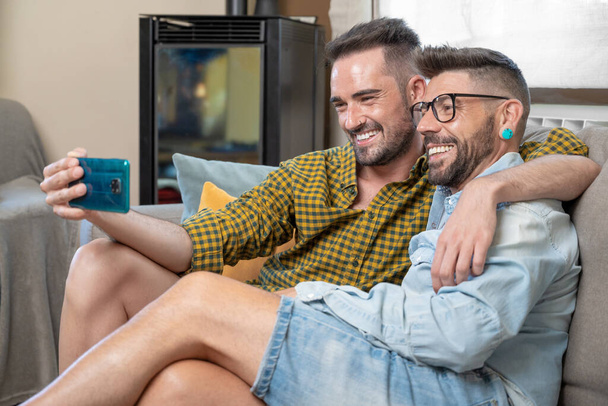 Gay Couple sitting in the sofa At Home Making Video Call On Mobile Phone. High quality photography. - Foto, immagini