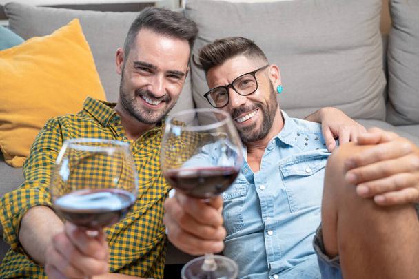 Gay couple in love sitting at home on the floor and toasting with red wine. High quality photography. - Fotografie, Obrázek