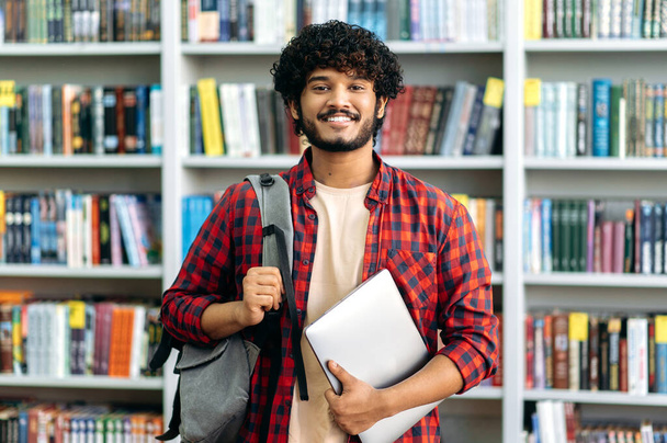Happy confident indian or arabian male student of university, in stylish casual wear, with backpack and laptop, stands in a library against the background of bookshelves, looks to the camera, smiles - Фото, зображення
