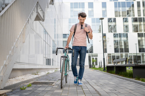 One man young adult male with brown hair and mustaches walking by the building with rucksack on his back and bicycle happy smile joyful real people copy space full length front view - Φωτογραφία, εικόνα