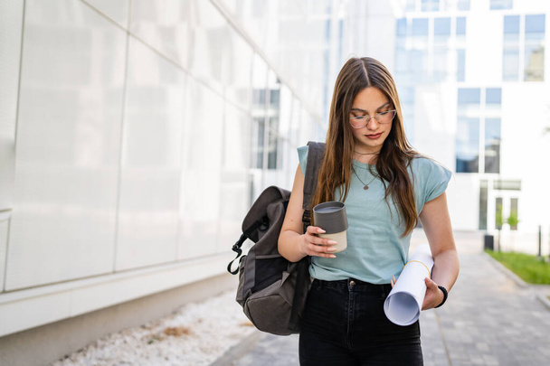 One woman young adult caucasian female student standing and walking in front of university with paper and cup of coffee outdoor in bright day real people copy space - Foto, afbeelding