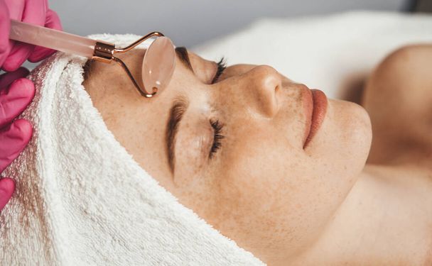 Client with her eyes closed receiving a facial massage with jade roller in the spa salon. Massage cabinet in the professional beauty clinic. Girl facial - Foto, Imagen