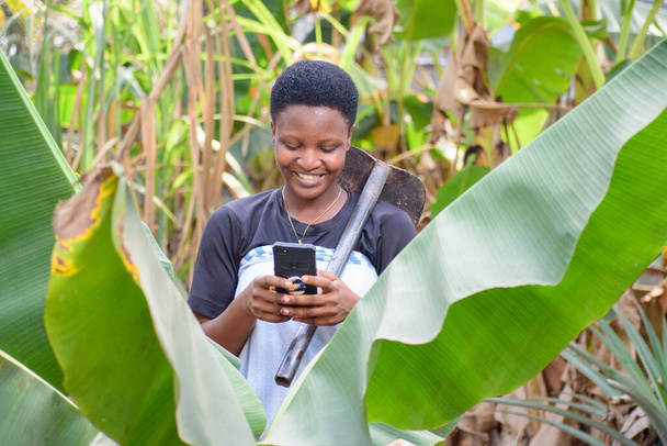Счастливая африканская женщина-фермер, деловая женщина или предприниматель, пользующаяся смартфоном на ферме, а также имеющая на плече фермерскую мотыгу - Фото, изображение