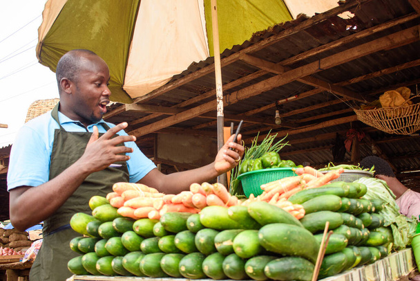 φωτογραφία αρχείου του αρσενικού αφρικανικού πωλητή παντοπωλείου με ποδιά, κατάπληκτος καθώς στέκεται και κοιτάζει επίμονα στο τηλέφωνό του - Φωτογραφία, εικόνα