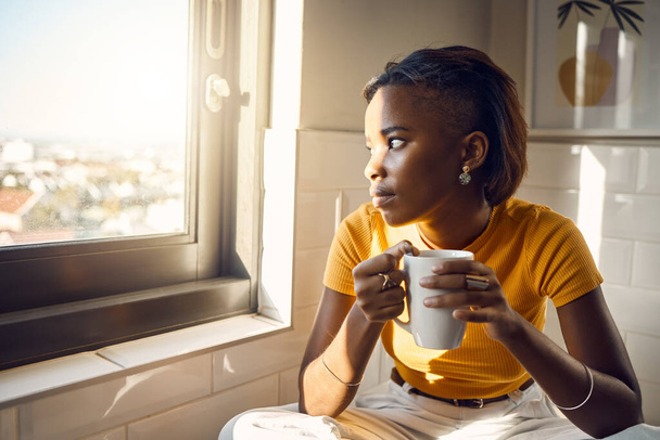 A thinking and coffee drinking black woman enjoys her morning routine while gazing outside a window on a bright sunny day. Beautiful female looks thoughtfully towards city while sitting at home. - Fotografie, Obrázek