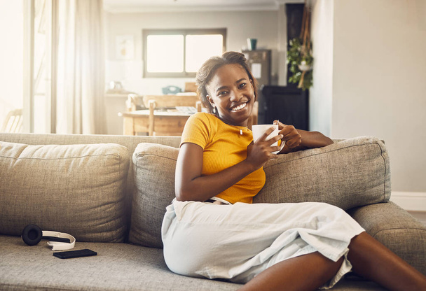 Beautiful, pretty and happy woman relaxing, drinking coffee and resting on the couch in the living room alone at home. Portrait of a smiling, cheerful and joyful black female enjoying a warm drink. - Fotografie, Obrázek