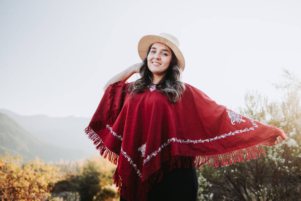 Усміхнена латинська традиційна жінка в червоному і жовтому пончо і капелюсі в природному просторі на заході сонця
. - Фото, зображення