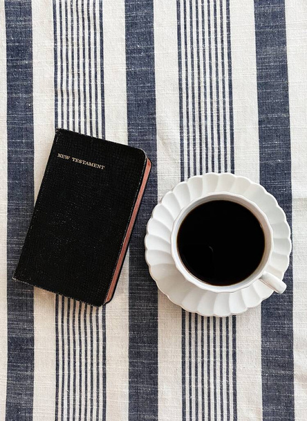A vertical shot of a pocket size Bible  and a cup of coffee on a stripe tablecloth surface - Фото, зображення