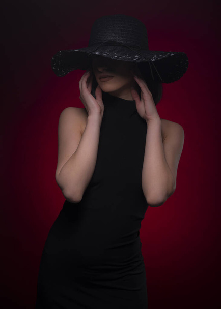Mystery attraktives Mädchen posiert auf rotem Hintergrund, während sie ihr Gesicht mit dem Hut versteckt - Foto, Bild