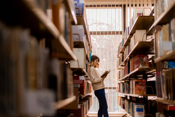 Beautiful girl reading a book while leaning on the bookshelves - 写真・画像