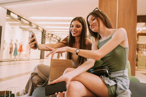 Two cute girls looking and laughing at the phone while sitting at the sofa in the mall - Foto, Imagen