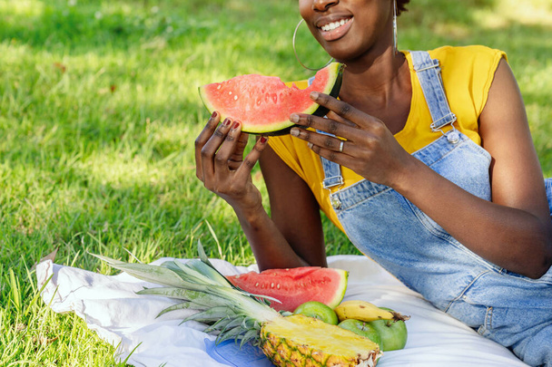 Short shot of an unrecognizable, very happy smiling black African woman lying in an outdoor park while eating a watermelon and more fruit at a picnic. High quality photo. - Foto, imagen