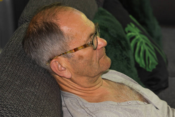 Side view of caucasian senior man with high temple in gray t-shirt and glasses falling asleep while watching TV on sofa. Evening leisure activity at home. Elderly life concept. - Photo, image