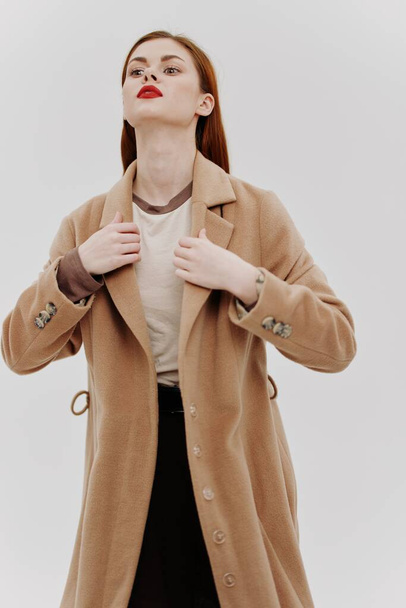Schieten voor de buitenkleding catalogus. Roodharige schoonheid in een beige full-length jas in de studio op een witte achtergrond. Hoge kwaliteit foto - Foto, afbeelding