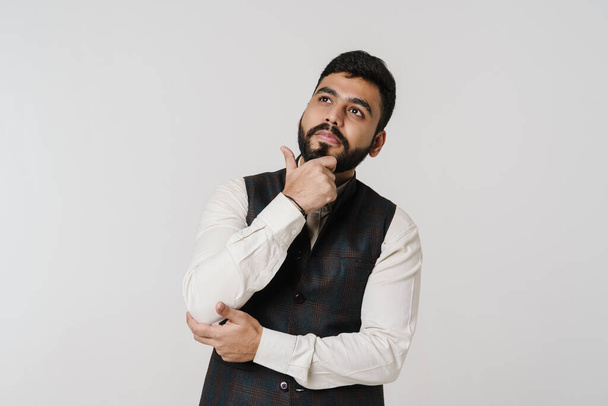 Bearded south asian man holding his chin and looking upward isolated over white wall - Foto, Imagem