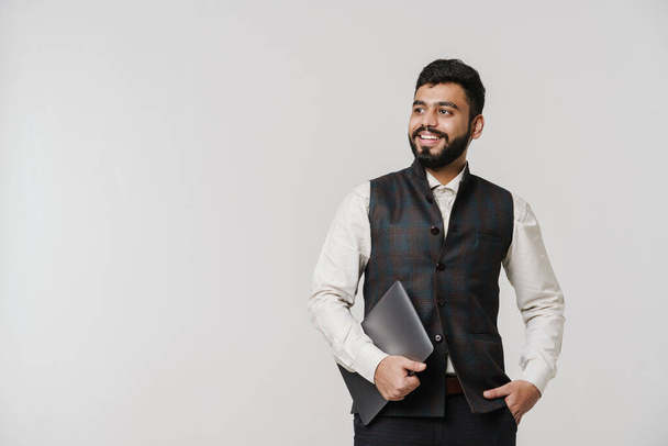 Bearded south asian man wearing vest laughing while posing with laptop isolated over white wall - Zdjęcie, obraz