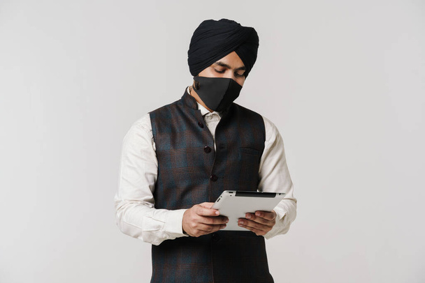 South asian man wearing turban and face mask using tablet computer isolated over white wall - Zdjęcie, obraz