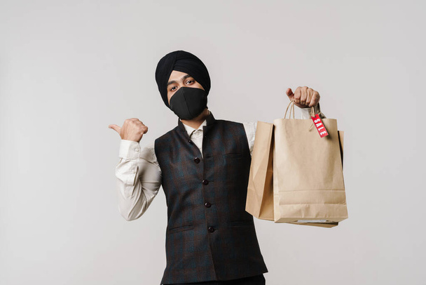 Yüzünde maske olan Güney Asyalı adam beyaz duvarın üzerinde izole edilmiş alışveriş torbaları tutarken parmağını kenarda tutuyor. - Fotoğraf, Görsel