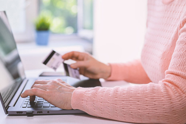 Mature woman works at home on a laptop. She is holding two credit cards in her hand. Closeup of hands, casual wear. Online shopping, work from home and freelance concept. - Foto, Imagen