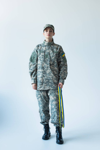 Повна довжина солдата, який тримає синьо-жовту стрічку і дивиться на камеру на сірому фоні
 - Фото, зображення
