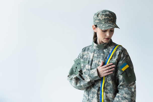 Young soldier in camouflage uniform with ukrainian flag touching blue and yellow ribbon isolated on grey  - Photo, Image
