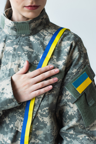 Cropped view of soldier with ukrainian flag on chevron touching blue and yellow ribbon isolated on grey  - Fotó, kép