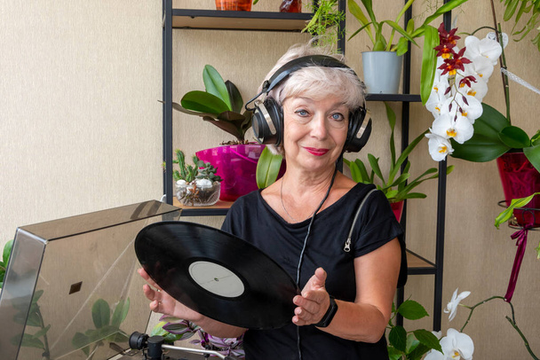 an elderly blonde woman 60-65 years old in old headphones looks at a vinyl record, a record player against the background of indoor flowers.  Concept: retirement vacation, audio therapy. - Fotoğraf, Görsel