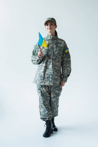 Smiling soldier in uniform holding ukrainian flag on grey background - 写真・画像