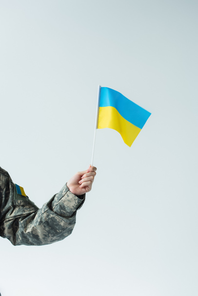 Cropped view of soldier in uniform holding ukrainian flag isolated on grey  - Фото, изображение