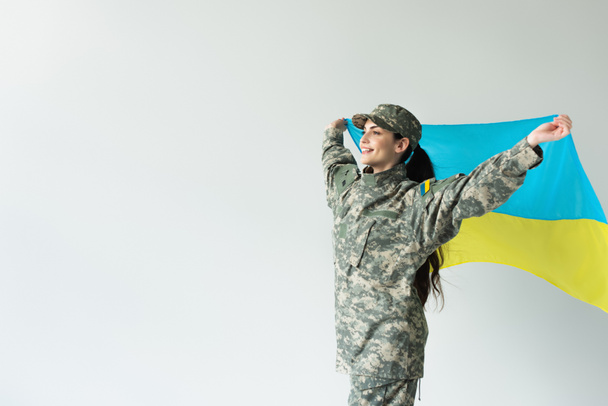 Happy servicewoman holding ukrainian flag isolated on grey  - Фото, изображение