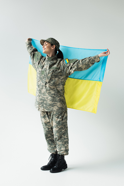 Side view of smiling soldier with closed eyes holding ukrainian flag on grey background - Fotó, kép