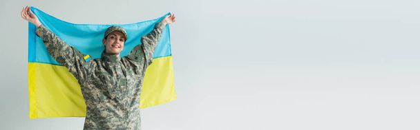 Boldog szolgálólány álcázó egyenruhában ukrán zászlót tart elszigetelve szürke zászlóval, zászlóval  - Fotó, kép