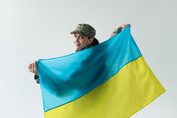 Smiling soldier looking at camera while holding ukrainian flag isolated on grey  - Fotó, kép