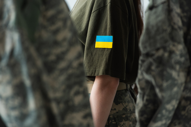 Cropped view of servicewoman with ukraining flag on chevron standing near soldiers isolated on grey  - Fotó, kép