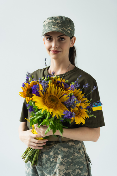Smiling soldier with ukrainian flag on chevron holding bouquet isolated on grey  - Foto, Imagen