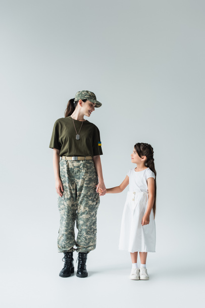 Side view of soldier with ukrainian flag on uniform holding hand of daughter on grey background - Foto, immagini