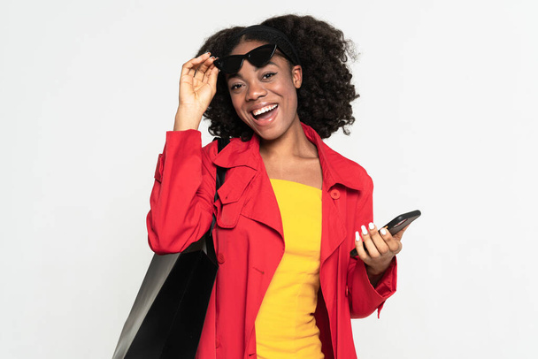 Young black woman using cellphone while posing with shopping bag isolated over white background - Fotografie, Obrázek