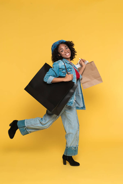 Young black woman laughing while walking with shopping bags isolated over yellow background - Valokuva, kuva