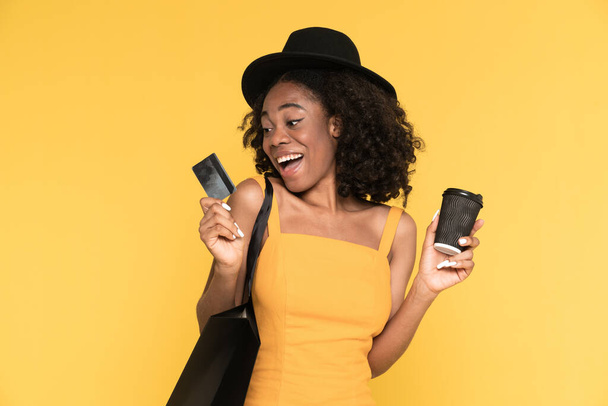 Black woman drinking coffee while posing with credit card and shopping bag isolated over yellow background - Фото, изображение